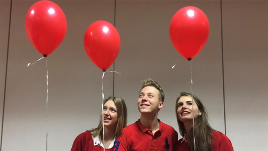Fotographie von zwei Schülersprecherinnen und einem Schülersprecher. Die drei halten jeweils einen roten Luftballon in der Hand und tragen passend dazu rote Polohemden. Das Bild stellt ein Beispiel für ein SMV-Projekt dar.