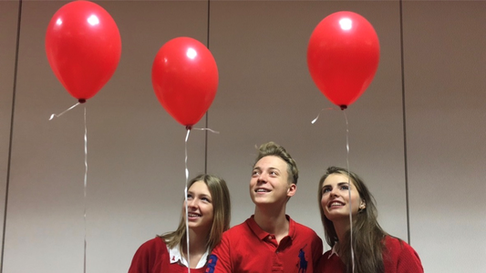 Fotographie von zwei Schülersprecherinnen und einem Schülersprecher. Die drei halten jeweils einen roten Luftballon in der Hand und tragen passend dazu rote Polohemden. Das Bild stellt ein Beispiel für ein SMV-Projekt dar. 